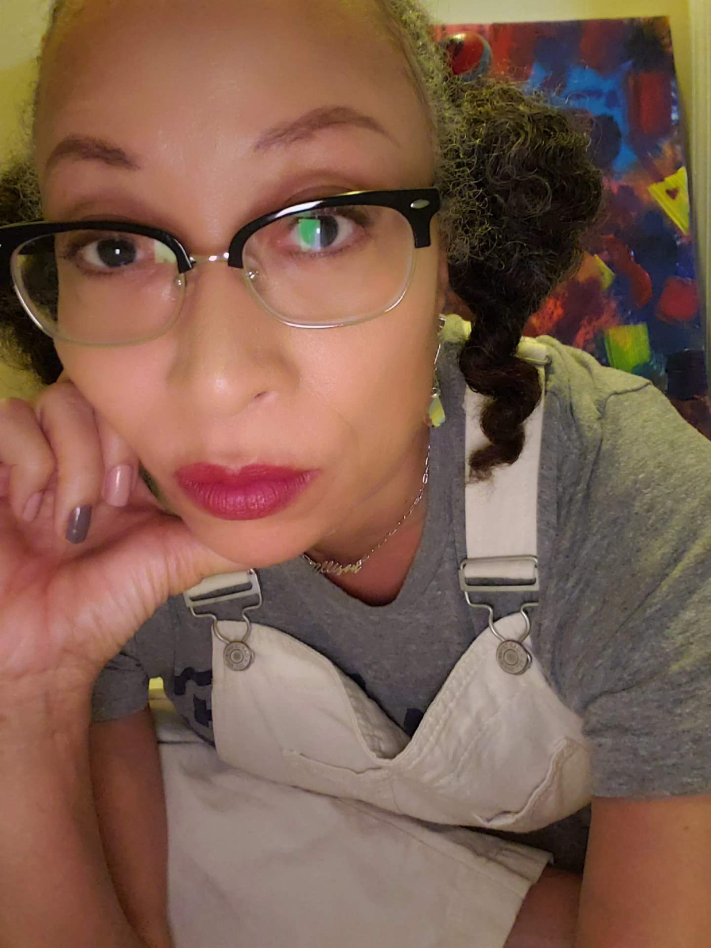 Allison Kalloo in her studio, chin resting on her hand, in glasses and white overalls in front of a colorful painting