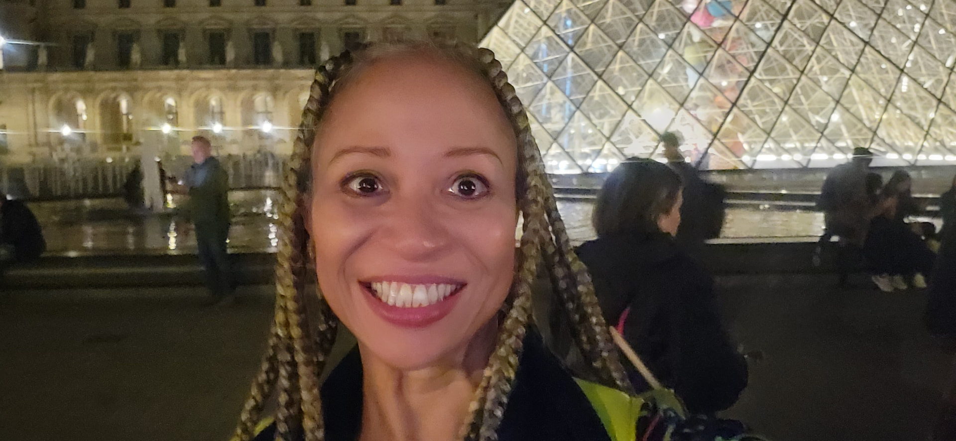 Allison grinning wide-eyed in front of the glowing Louvre pyramid at night
