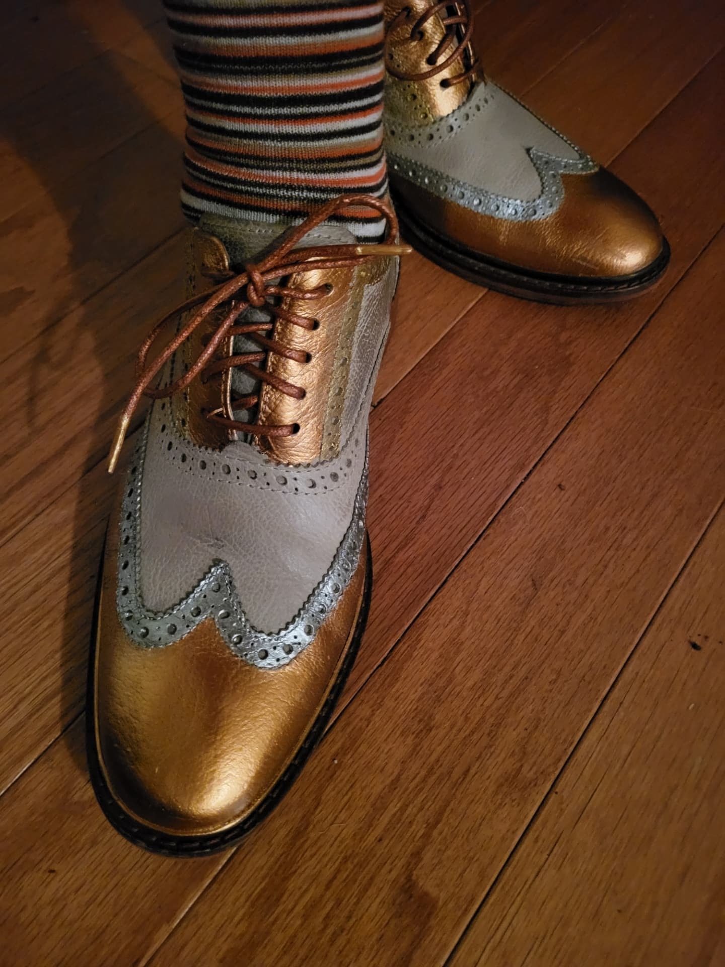 Overhead candid photograph of copper and grey wingtip brogues with striped socks on a warm wood floor