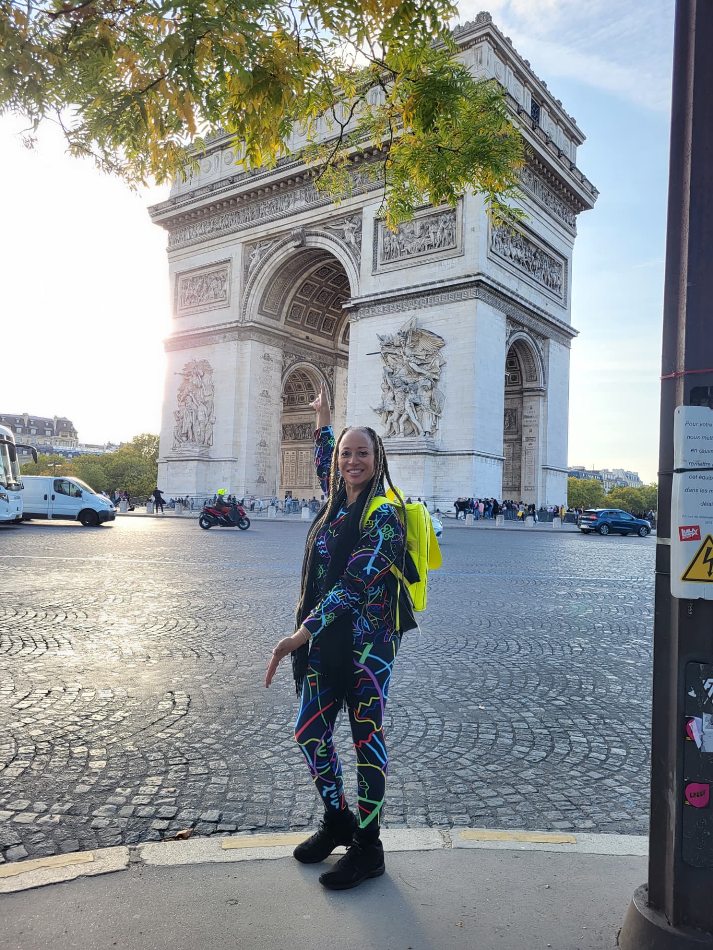 Allison in a bright patterned outfit and neon backpack pointing up at the Arc de Triomphe