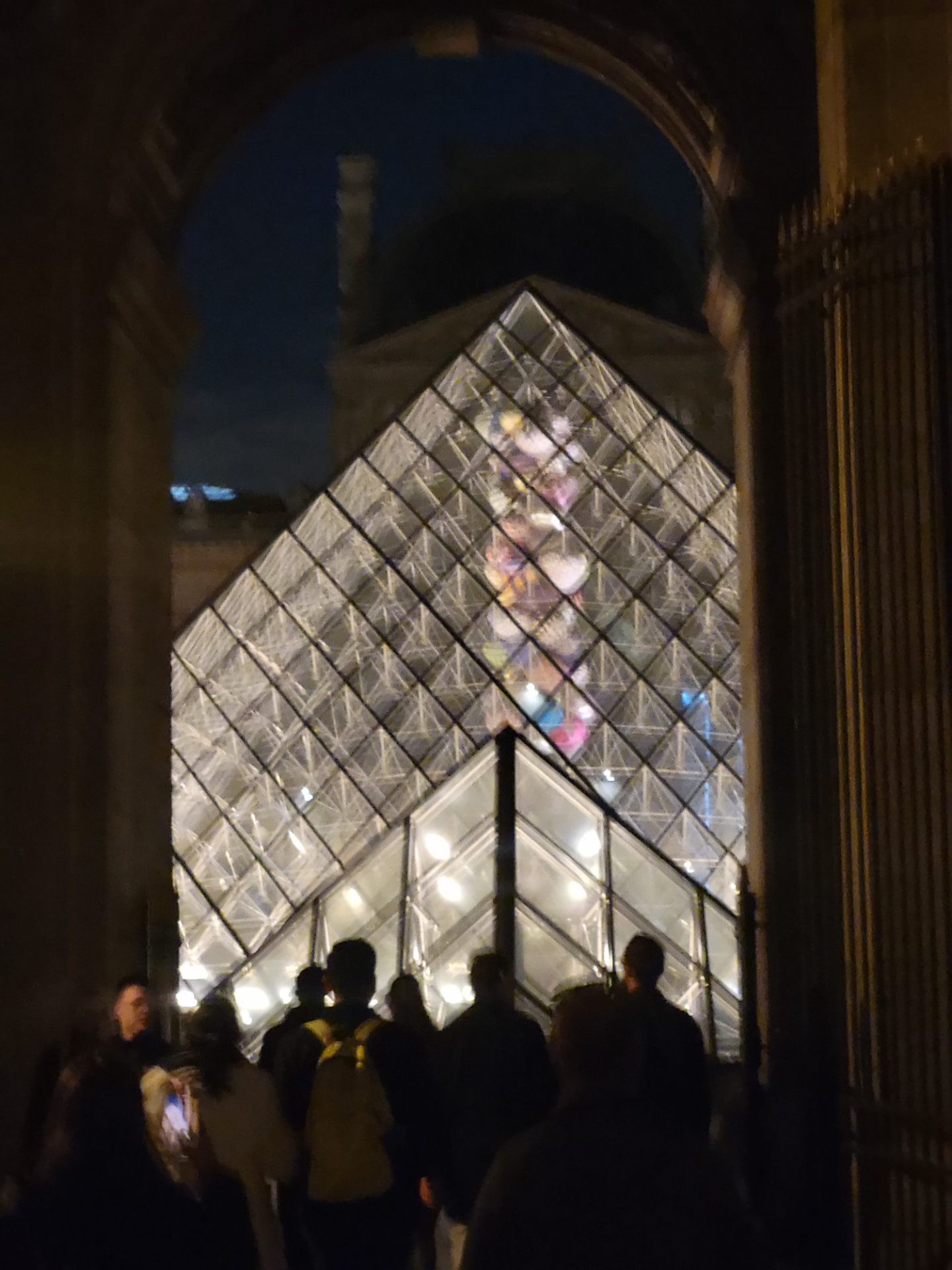 The Louvre pyramid glimpsed through a dark stone archway, visitors silhouetted below