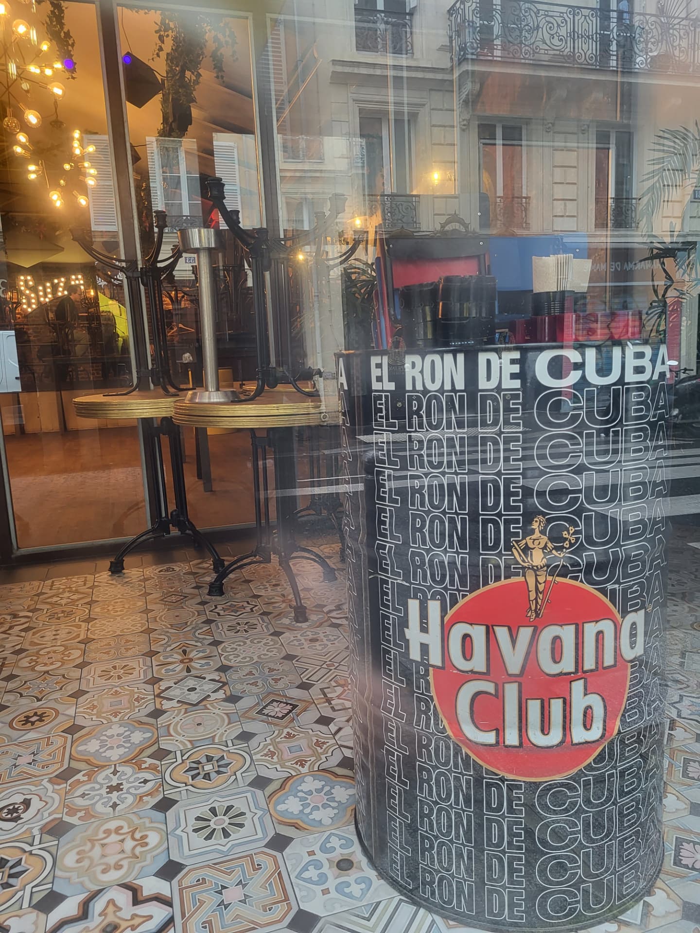 A Paris café window with a Havana Club rum barrel, patterned tile floor and reflected balconies