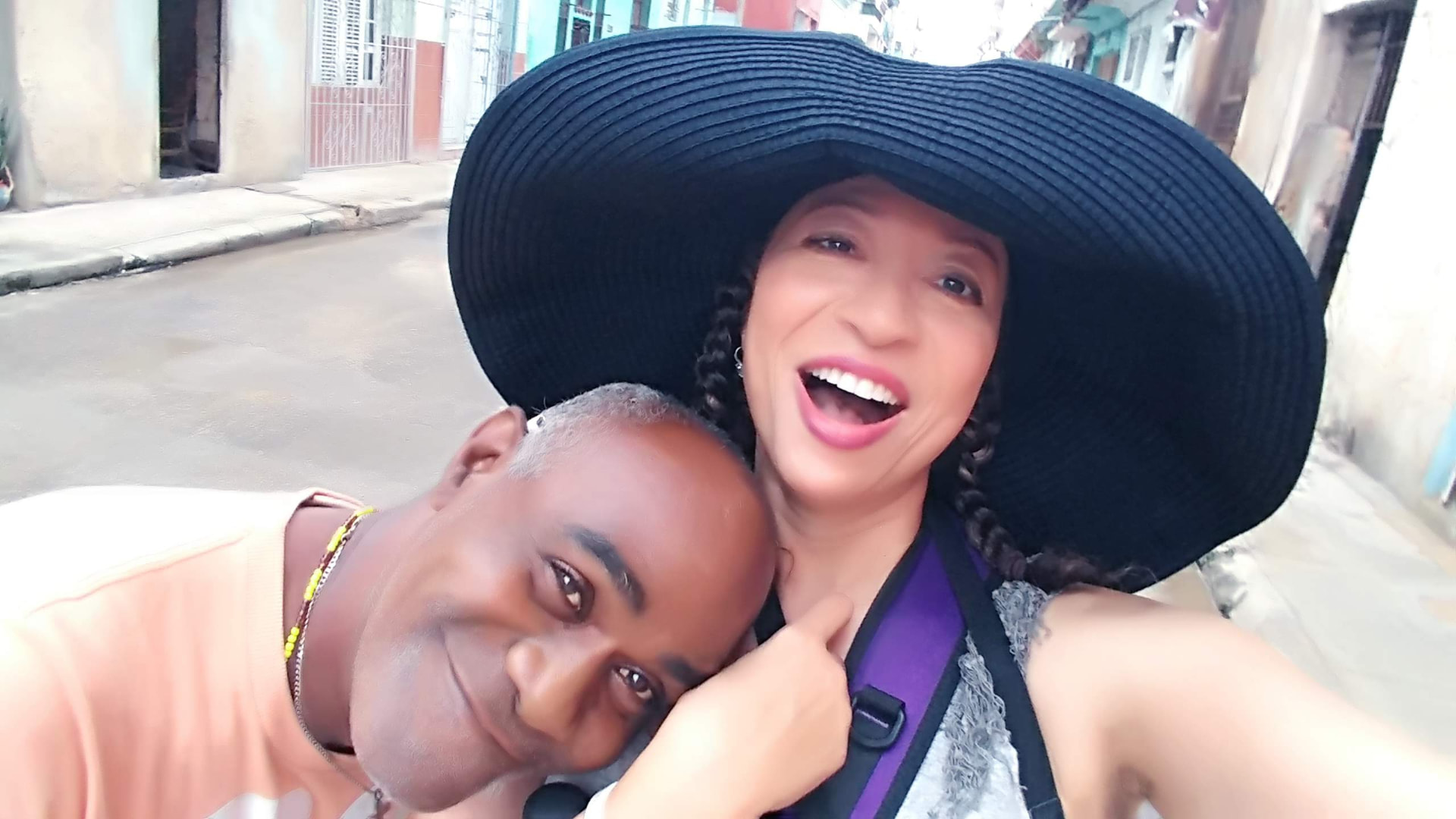 Allison laughing on a Havana street in a wide black hat beside a companion